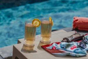 Two drinks and a bottle of sunscreen with swimming pool in the background